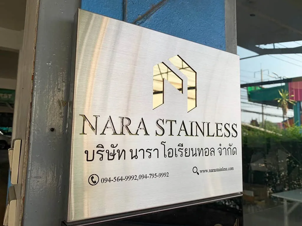 Shiny stainless steel sign with gold-colored accents showcasing NARA Stainless logo and Thai text, mounted outdoors with a reflection of the surrounding environment.