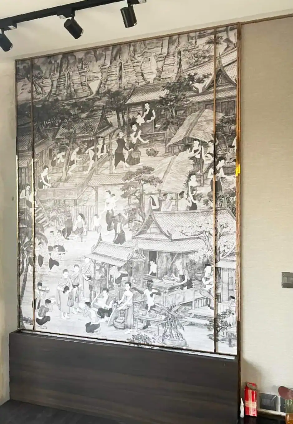 Intricate black and white mural depicting traditional Thai village life with houses, trees, and people engaging in daily activities, framed by a decorative brass stainless steel trim.