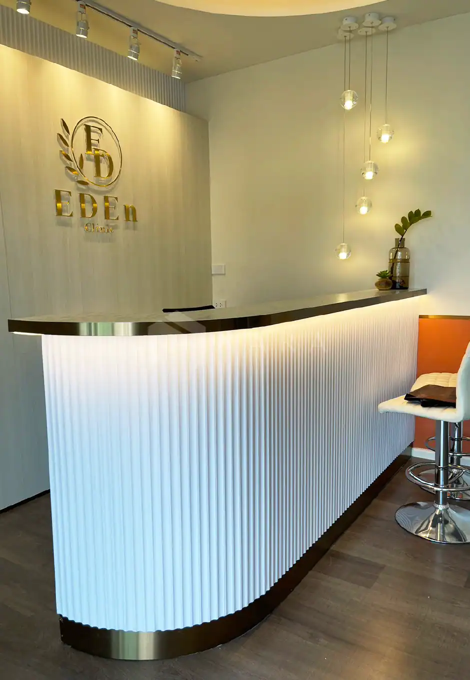 Shiny gold-colored stainless steel trim accents the modern white reception desk with a curved design and vertical ribbing, creating an elegant and welcoming office entrance with pendant lighting and minimalist decor.