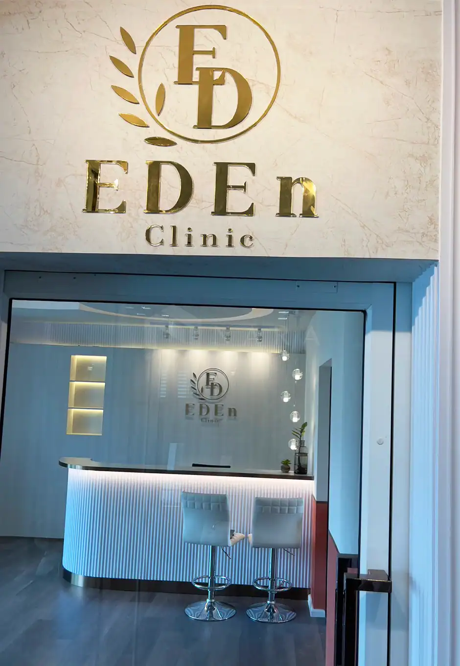 Shiny gold stainless steel frame and signage at EDEN Clinic entrance, featuring decorative stainless steel trim and modern interior with illuminated reception desk and cozy seating area, creating an elegant and inviting atmosphere.