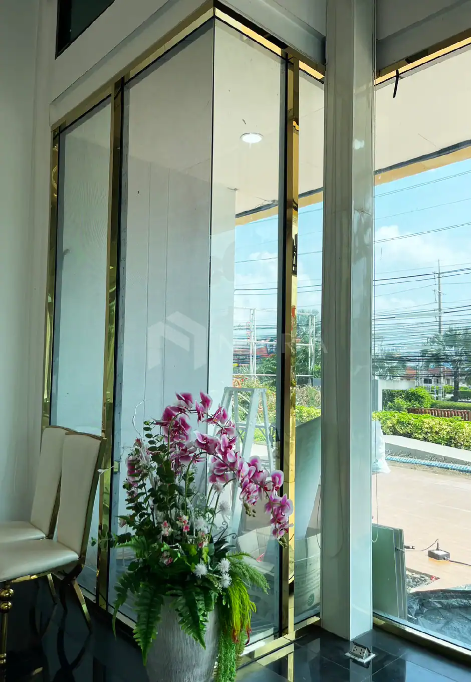 Shiny gold-colored stainless steel decorative corner trim inside a modern building with large glass windows and floral arrangements.