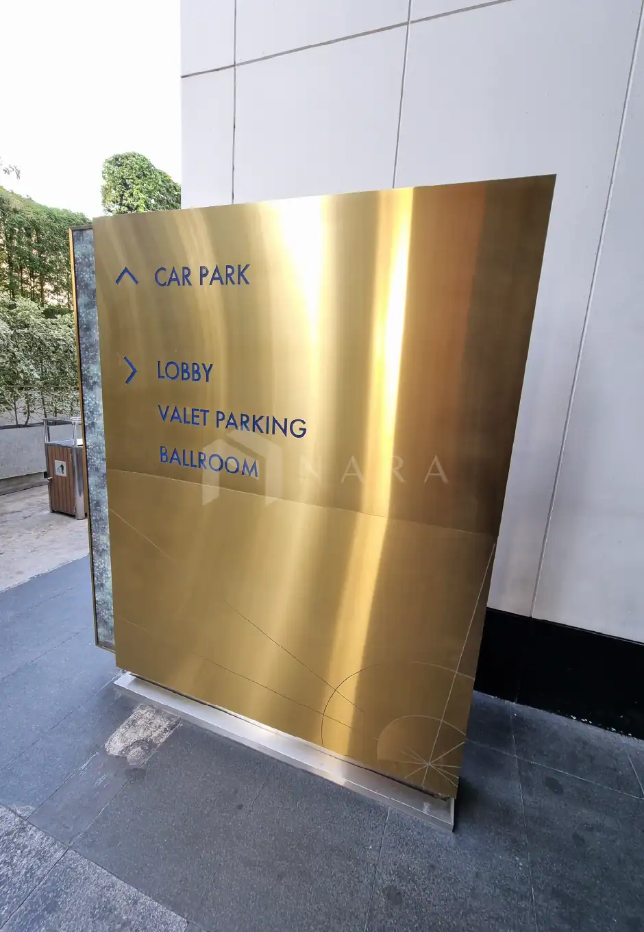 Shiny gold stainless steel directory sign with engraved blue text, providing directions to car park, lobby, valet parking, and ballroom at a modern building.