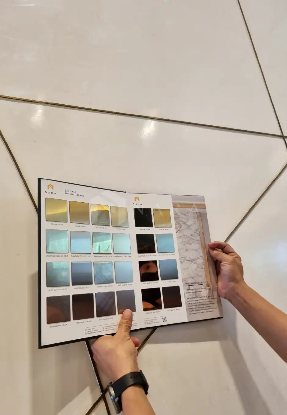 Shiny gold stainless steel trim sample sheet on white tiled floor, showcasing various stainless steel color options with hands holding the sample for home or commercial interior design.
