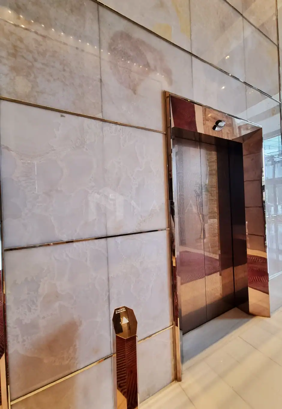 Shiny gold stainless steel elevator frame with beige tiled wall accents, showcasing luxurious interior design featuring stainless steel decorative trims and corner details for a modern upscale look.