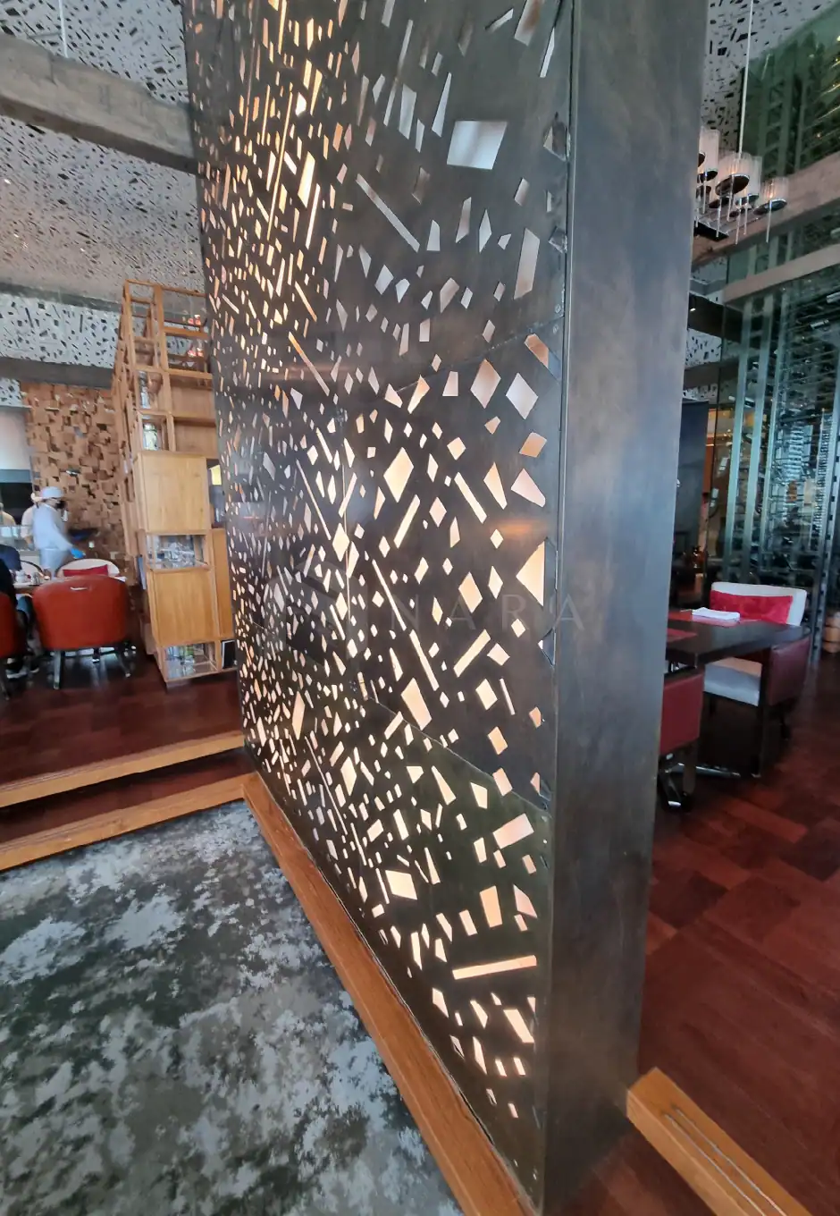 Vivid image of a decorative stainless steel partition with intricate geometric cutouts, illuminated from behind, enhancing modern interior aesthetics in a stylish restaurant setting.