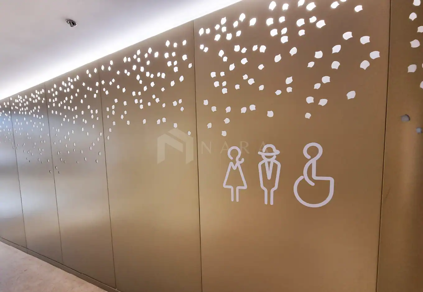 Shiny gold stainless steel wall with decorative cutouts and signage for restrooms, including accessible facilities, showcasing modern stainless steel design and craftsmanship.